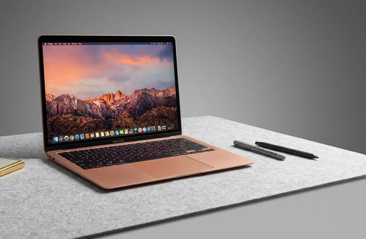 Refurbished Apple MacBook on a gray surface, with a scenic mountain wallpaper and pens nearby.