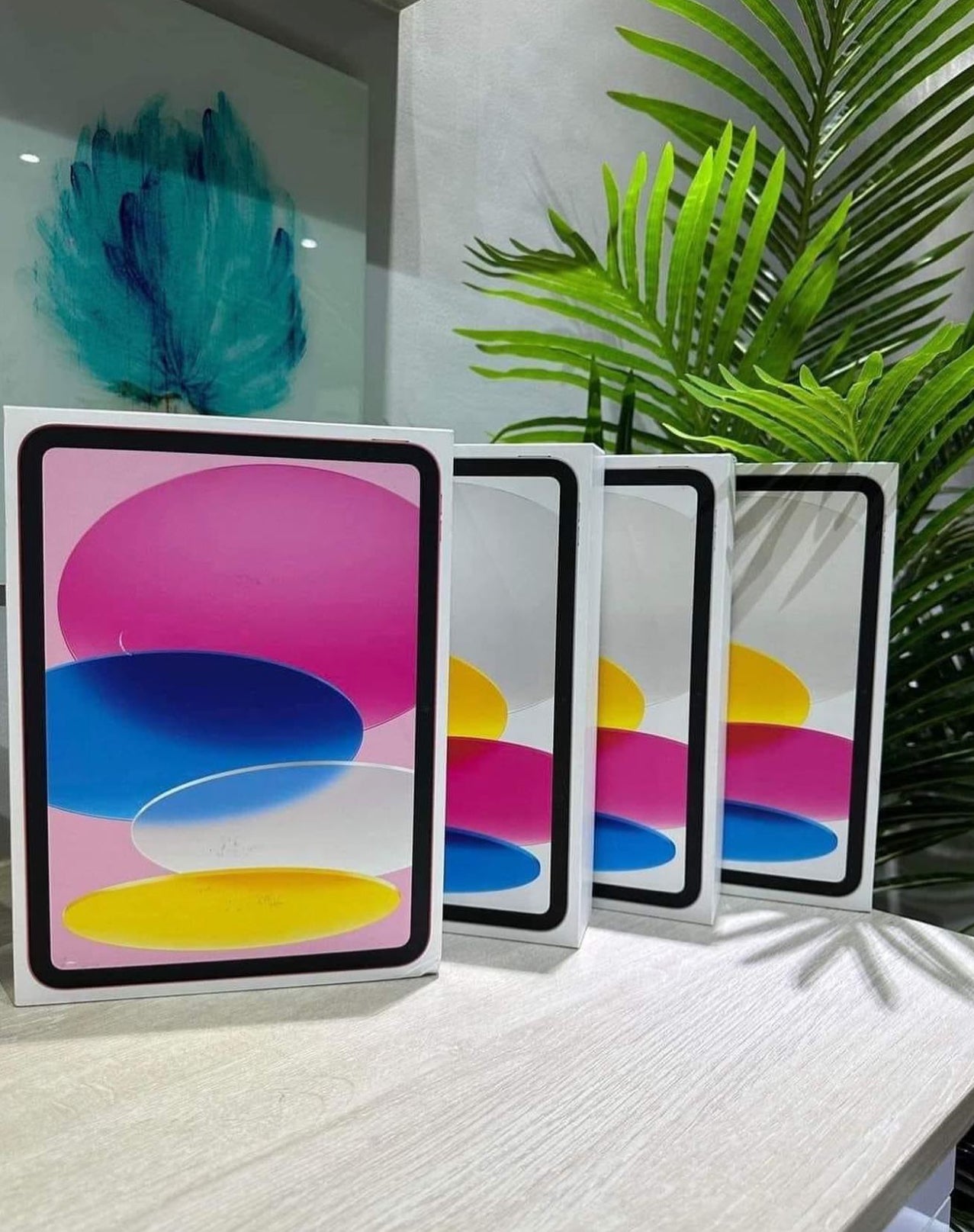 Four refurbished Apple devices with colorful abstract designs displayed on a table next to a green plant.